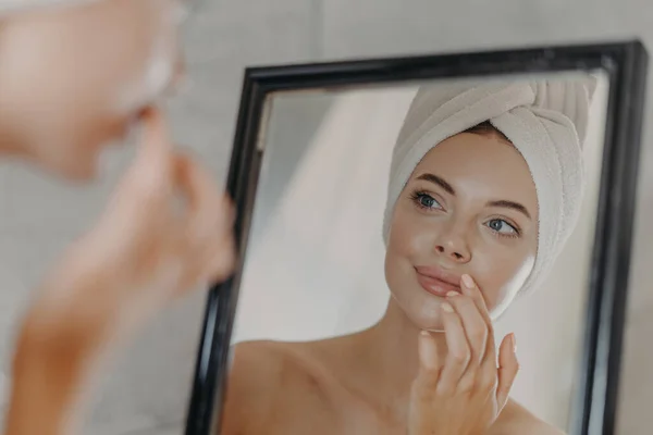 健康的女人只穿少量化妆品 照顾皮肤和嘴唇 照镜子 站在光秃秃的肩膀上 头上戴浴巾 拥有健康完美的肌肤 卫生概念 — 图库照片