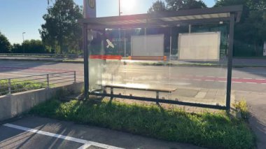 Boş bir otobüs durağının sakin manzarası yeşilliklerle çevrili, sabah güneşiyle aydınlatılmış, modern tasarım ve şehir ulaşımı sergileniyor..