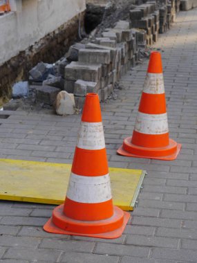 İki turuncu ve beyaz trafik konisi tuğla kaldırımda. Külahlar sarı bir tahtaya konur.