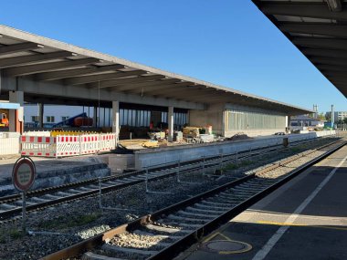A view of a train station platform under construction, showcasing rail tracks and building renovations in bright daylight.
