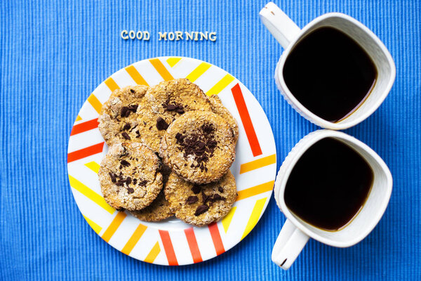 Овсяное печенье с шоколадом на голубой салфетке и двумя чашками кофе, надпись с добрым утром
