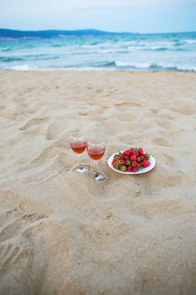 two glasses of wine and a plate of strawberries on a sandy beach, a picnic near the sea