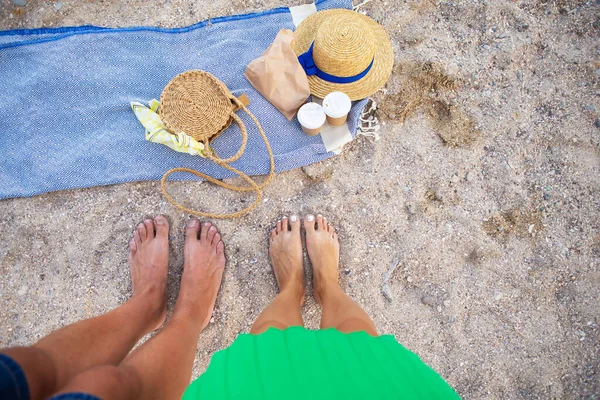 Picnic near the sea. A blue plaid on which are glasses with coffee, a bag, a hat. Feet of a couple in love. View from above.