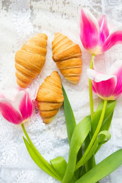 Drei Croissants und leuchtend rosa Tulpen auf Spitzentischdecke — Stockfoto