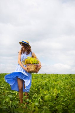 Yeşil bir tarlada elinde bir sepet çiçekle duran şapkalı bir kız..