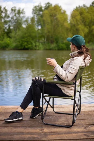 Rıhtımda oturan, gölde balık tutan, gülümseyen, dikiz manzaralı bir kız. Açık hava aktiviteleri, güzel bir yaz günü. Kız kahve içiyor.