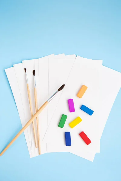 Le vernici multicolori, le spazzole ad acquerello di dimensioni diverse si trovano insieme con carta ad acquerello su uno sfondo blu. Lezioni di disegno. Vista dall'alto. — Foto Stock
