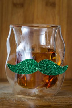 Close up of a glass of whiskey with a green mustache over a wooden background as a concept for Saint Patricks day clipart