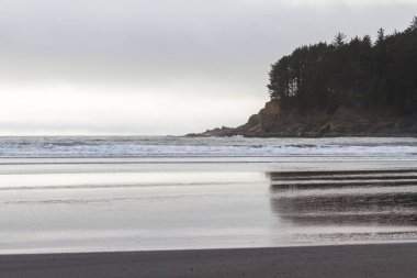 Oregon sahilinde karamsar bir öğleden sonra. Düşük yoğunluklu bulutlar hafif yamalar arasından geliyor ve suya yansıyor.