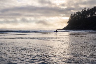 Okyanustan çıkıp sahilde yürüyen bir adam elinde sörf tahtasıyla suya yansıyan bulutlardan gelen altın ışıkla.