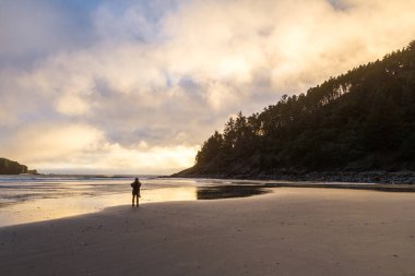 Avcılardan güzel bir gün batımının tadını çıkaran bir kadın. Parlak bulutlar ıslak kumu yansıtıyor.