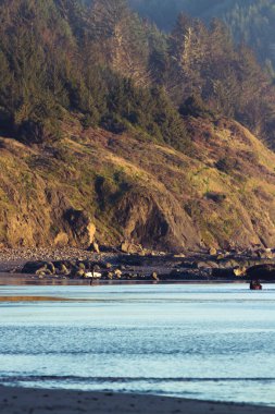 Güneş ışığı arkadaki dağı aydınlatırken kumda sörf tahtası taşıyan bir adam kuma parlak bir yansıma ekliyor.