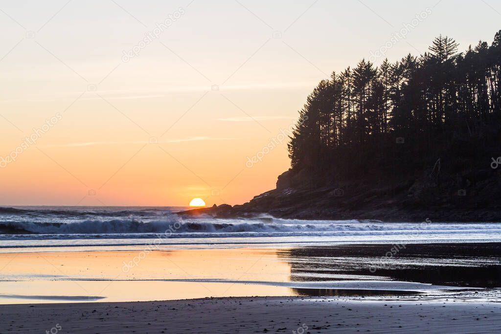 Glowing orange sun setting behind the horizon adding an orange glow to the sky and a reflection on the wet sand in Hunters Cove a popular surfing spot in the Oregon coast
