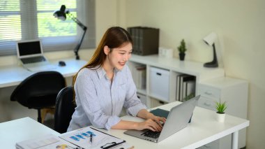 Ev ofisinin bilgisayarında çalışan gülümseyen genç bir kadın. Masasında belgeler var..