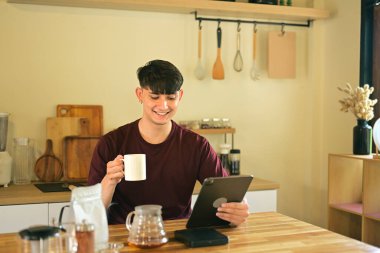 Elinde kahve fincanı olan gülümseyen beyaz bir adam mutfakta dijital tablet üzerinde geziniyor..