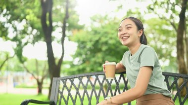 Gülümseyen genç bir kadın, elinde kahve fincanı tutarken bankta dinleniyor..