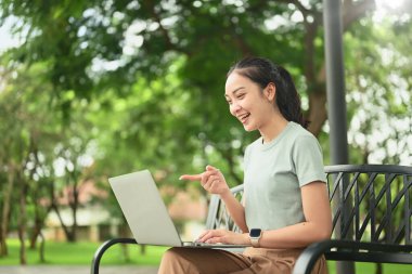 Gülümseyen genç bir kadın, dışarıda parkta otururken bilgisayarında bir video görüşmesi yapıyor..