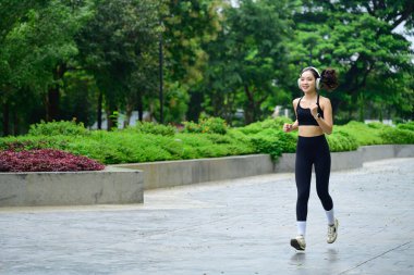 Çevresi yeşilliklerle çevrili yolda koşup müzik dinleyen formda bir bayan sporcu..