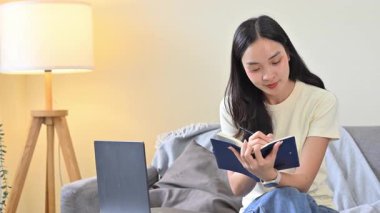 Gülümseyen kadın çevrimiçi öğrenme sırasında dizüstü bilgisayara bakarken not alıyor..