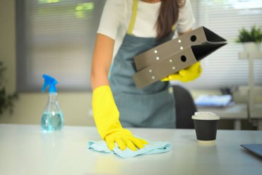 Önlük ve eldiven giyen kadın işçi modern ofiste sprey şişesi ve kumaşla masaları siliyor..