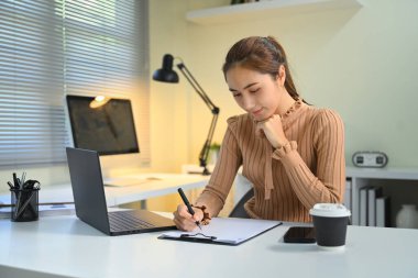 Dizüstü bilgisayarla çalışırken panoya notlar yazan odaklanmış genç bir kadın..