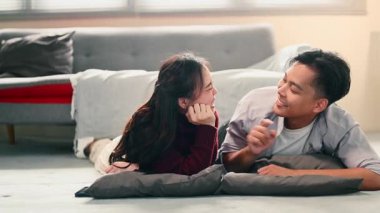 Yastıklarla yerde yatan mutlu çift, evde birbirlerine sevgiyle bakıyor ve gülümsüyor..