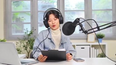 Evde mikrofon, dizüstü bilgisayar ve kulaklıkla podcast yapan genç bir kadın..