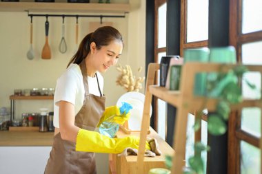 Mutfak raflarını sprey şişesi ve bezle temizlerken gülümseyen genç bir kadın..