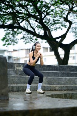 Açık hava kardiyo seansı sırasında çömelme egzersizi yapan gülümseyen bir sporcu..