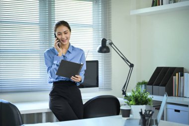 Modern ofisteki belgeleri incelerken akıllı telefondan konuşan gülümseyen iş kadını..