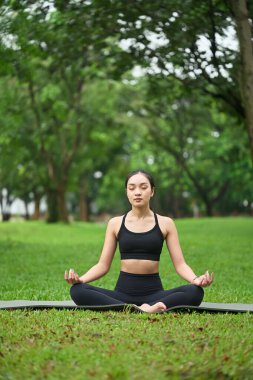 Nilüfer pozisyonunda meditasyon yapan genç bir kadın huzur dolu yeşil bir parkta yoga minderi üzerinde..