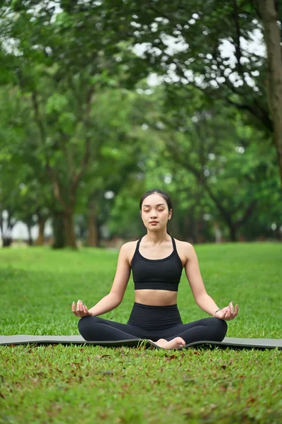 Nilüfer pozisyonunda meditasyon yapan genç bir kadın huzur dolu yeşil bir parkta yoga minderi üzerinde..