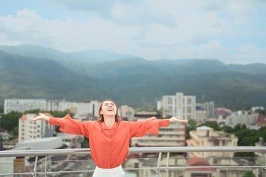 Neşeli bir kadın kolları açık çatıda duruyor, neşeyle gülümsüyor şehir manzarasına ve dağ manzarasına karşı..