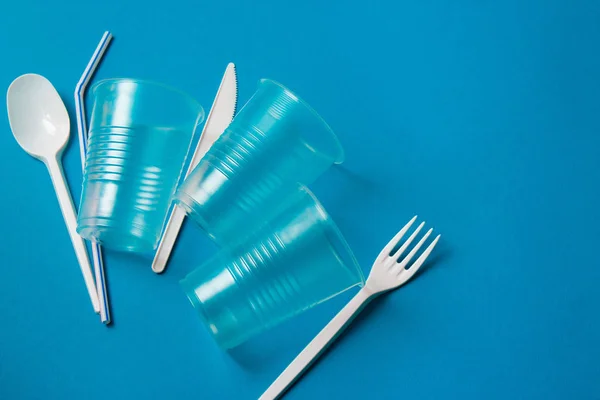 White single-use plastic knife, spoon, fork and plastic drink straws on a blue background. Say no to single use plastic. Environmental, pollution concept.