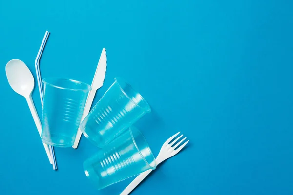 White single-use plastic knife, spoon, fork and plastic drink straws on a blue background. Say no to single use plastic. Environmental, pollution concept.