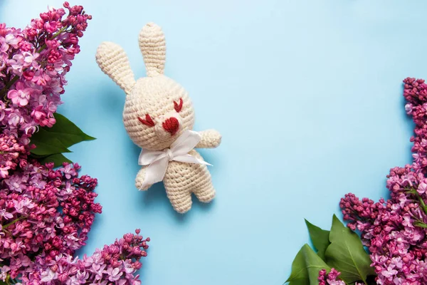 Lilac flowers with knitting toy rabbit on blue background. Top view, flat lay, copy space.