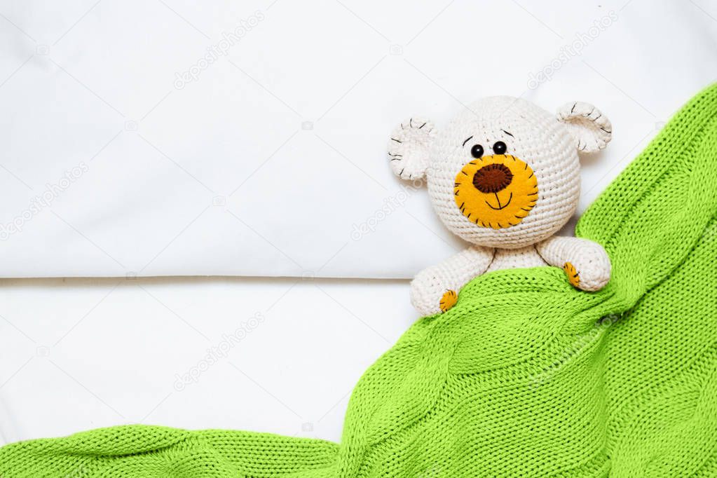 A small knitted amigurumi baby toy-bear is covered with a green blanket, flat lay, top view