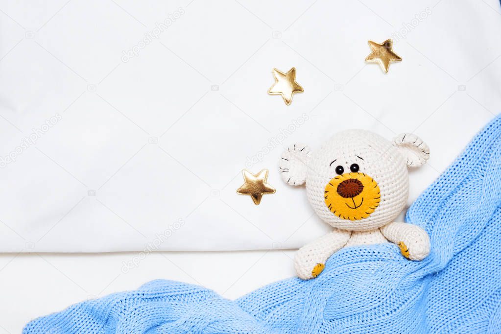 A small knitted amigurumi baby toy-bear is covered with a blue blanket, flat lay, top view