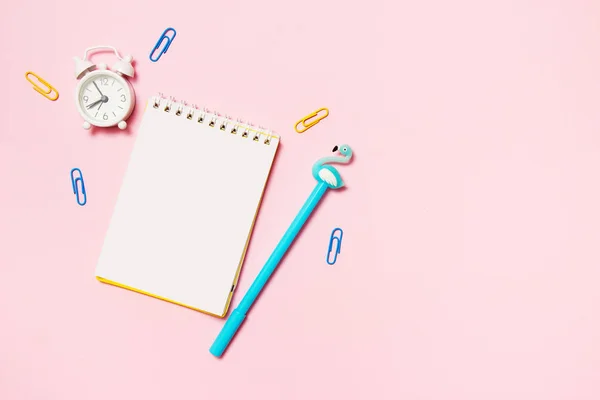 Блокнот с цветной ручкой, скрепками и будильником на розовом фоне. Школа, рабочее пространство, крайний срок. Плоский, вид сверху — стоковое фото