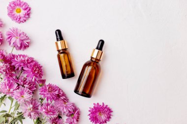 Damlatan cam şişelerden oluşan düz bir döşeme. Cilt, çiçek kasımpatıları eşliğinde beyaz arka planda asgari düzeyde taklit edilebilecek temel yağ ürünleri.