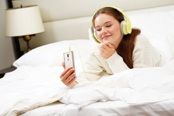 携帯電話スマート フォンの顔がヘッドフォンで音楽を聴いて夢を見て笑みを浮かべて見ていると白いベッドで美しい魅力的な若い白人女性ブルネット朝 — ストック写真