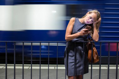 Güzel, genç bayan banliyö çalışanı, modern bir tren istasyonunda günlük trenini beklerken cep telefonunu kullanıyor.)