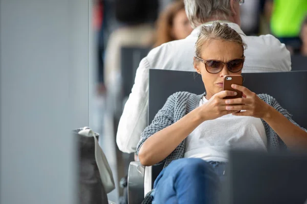 国際空港の出発ゲートで飛行機に搭乗を待っている間彼女の携帯電話を使用してカジュアルな金髪の若い女性 — ストック写真