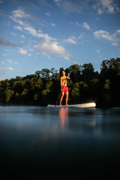 一个英俊的小伙子在桨板上 在温暖的黄昏阳光下 在一条可爱的河上做一个伟大的运动 — 图库照片