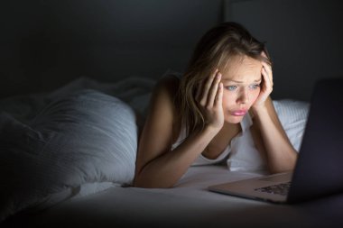 Genç ve güzel kadın yatakta onun laptopunda korkunç/üzücü bir şey karanlık bir odada (sığ Dof; renk tonlu görüntü geceleri izlerken)