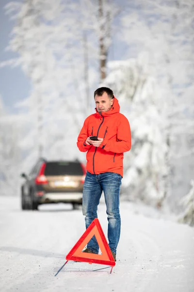 在寒冷的冬日里 一个英俊的男人在他的车子在荒无人烟的地方抛锚后 设置了一个三角形的警告 并呼救 — 图库照片