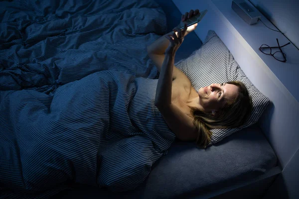 Wanita cantik setengah baya menggunakan ponselnya di tempat tidur di rumah. — Stok Foto