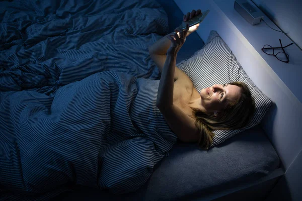 Pretty, middle-aged woman using her cell phone in bed at home — Stock Photo, Image