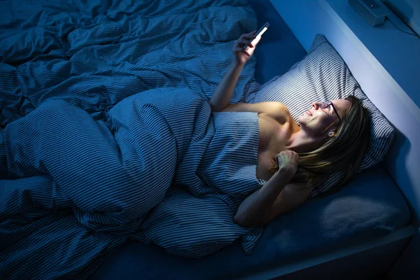 Красивая женщина средних лет использует свой мобильный телефон в постели дома — стоковое фото