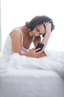 Güzel genç bir kadın sabah ilk iş olarak akıllı telefonunu kullanarak uyuyor.